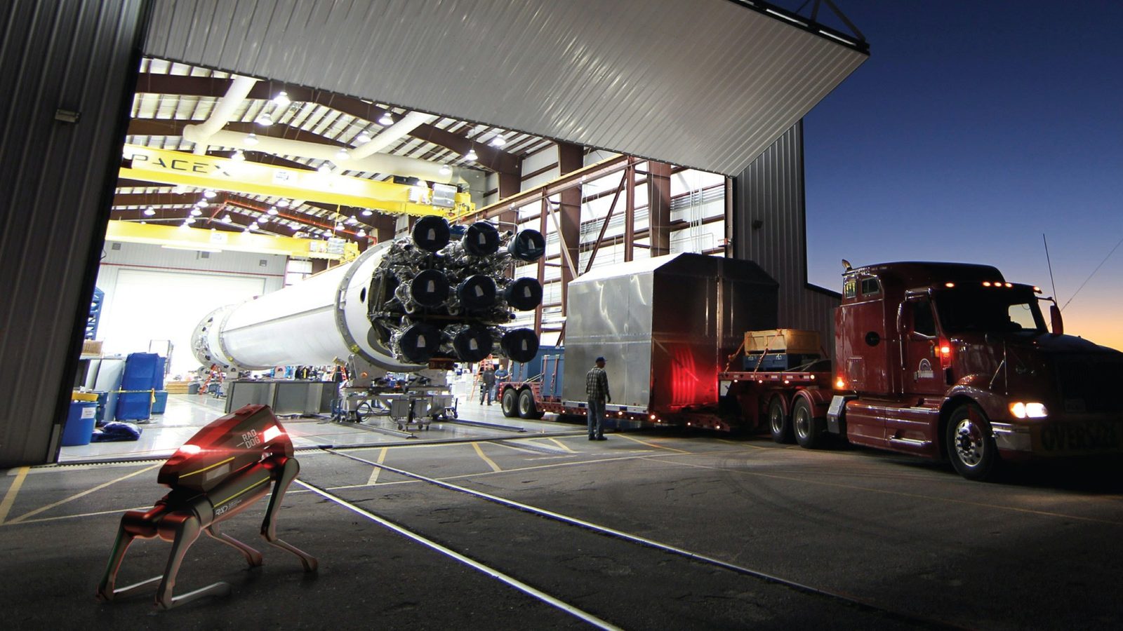 RADDOG at a rocket facility with a Falcon 9 booster and transport truck at dusk